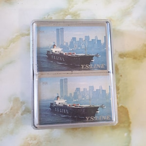 Baraja de cartas de dos barajas, década de 1970. Yamashita Shinnihon Steam Ship Co. Ltd. Diseño de la foto de un barco y las Torres Gemelas de Nueva York. En estuche de plástico.