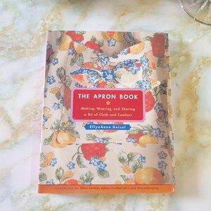 May include: A hardcover book titled "THE APRON BOOK" with a colorful fruit and floral pattern. The cover features a red rectangle with white text and the author's name. The book is about making, wearing, and sharing aprons.