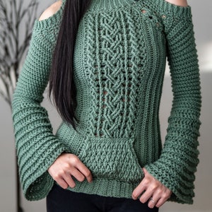 TCDDIY crochet pattern of a cold shoulder bell sleeve hoodie featuring intricate cable stitch detail on the body with alpine stitch hoodie pockets made in light sage yarn.