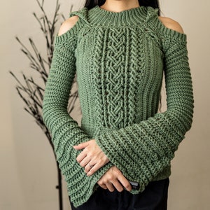 TCDDIY crochet pattern of a cold shoulder bell sleeve hoodie featuring intricate cable stitch detail on the body with alpine stitch hoodie pockets made in light sage yarn.