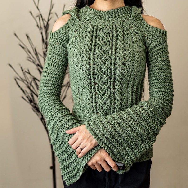 TCDDIY crochet pattern of a cold shoulder bell sleeve hoodie featuring intricate cable stitch detail on the body with alpine stitch hoodie pockets made in light sage yarn.