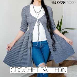 Pode incluir: Um cardigan cinza de crochê com um design de saia rodada. O cardigan tem um fecho de laço e mangas três quartos. O texto "CROCHET PATTERN" é exibido na parte inferior da imagem. O cardigan é usado sobre uma blusa branca e jeans azul.