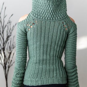 TCDDIY crochet pattern of a cold shoulder bell sleeve hoodie featuring intricate cable stitch detail with alpine stitch hoodie pockets made in light sage yarn, back view.