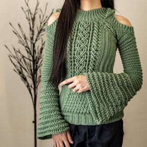 TCDDIY crochet pattern of a cold shoulder bell sleeve hoodie featuring intricate cable stitch detail on the body with alpine stitch hoodie pockets made in light sage yarn.