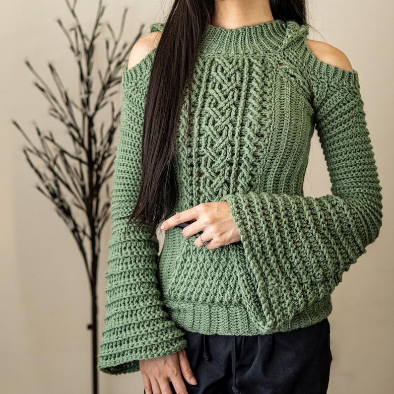 TCDDIY crochet pattern of a cold shoulder bell sleeve hoodie featuring intricate cable stitch detail on the body with alpine stitch hoodie pockets made in light sage yarn.