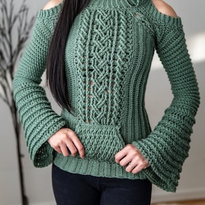 TCDDIY crochet pattern of a cold shoulder bell sleeve hoodie featuring intricate cable stitch detail on the body with alpine stitch hoodie pockets made in light sage yarn.