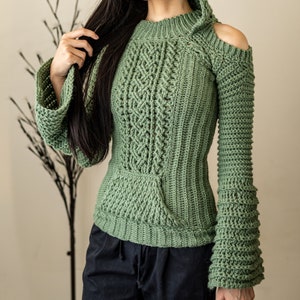 TCDDIY crochet pattern of a cold shoulder bell sleeve hoodie featuring intricate cable stitch detail on the body with alpine stitch hoodie pockets made in light sage yarn.
