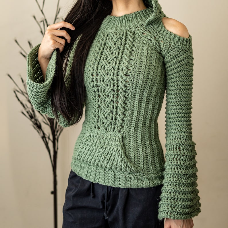 TCDDIY crochet pattern of a cold shoulder bell sleeve hoodie featuring intricate cable stitch detail on the body with alpine stitch hoodie pockets made in light sage yarn.