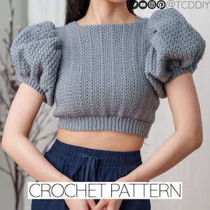 May include: A gray crochet crop top with puffed sleeves. The top is a short, fitted style with a round neckline. CROCHET PATTERN is written in black text at the bottom of the image.