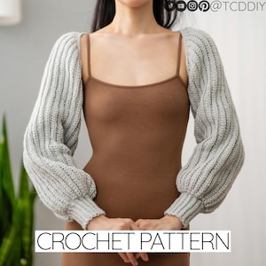 May include: A close-up of a person wearing a grey crochet shrug over a brown tank top. The shrug has long sleeves and a loose fit. The text "CROCHET PATTERN" is at the bottom of the image.