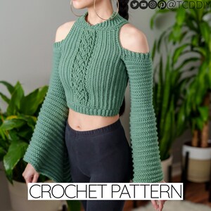 Può includere: Un top corto di uncinetto verde con un motivo a treccia e maniche a campana. Il top ha degli spacchi sulle spalle.