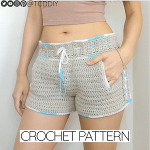 May include: Crochet shorts pattern. The shorts are beige with white and light blue trim. The shorts have a drawstring waist.
