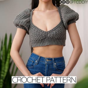 Könnte beinhalten: Ein graues Häkel-Crop-Top mit Puffärmeln. Das Top hat einen tiefen V-Ausschnitt und ein Häkelmuster. CROCHET PATTERN steht in weißer Schrift am unteren Rand des Bildes.