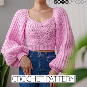 May include: A pink crochet top with a sweetheart neckline and long, puffy sleeves. The top is cropped and has a cable knit pattern. The model is wearing blue jeans.