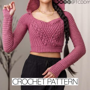 May include: A pink crocheted crop top with long sleeves and a hood. The top features a cable knit pattern and a sweetheart neckline. The text "CROCHET PATTERN" is visible at the bottom of the image.