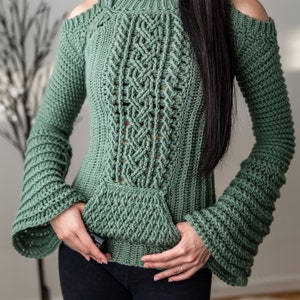 TCDDIY crochet pattern of a cold shoulder bell sleeve hoodie featuring intricate cable stitch detail on the body with alpine stitch hoodie pockets made in light sage yarn.