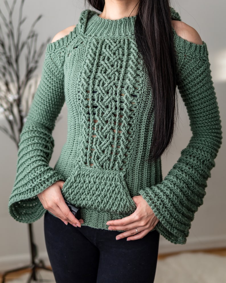 TCDDIY crochet pattern of a cold shoulder bell sleeve hoodie featuring intricate cable stitch detail on the body with alpine stitch hoodie pockets made in light sage yarn.