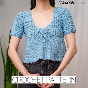 May include: A light blue crochet top with short sleeves and a V-neckline. The top features a tie detail at the bust and is paired with black jeans. The words "CROCHET PATTERN" are displayed at the bottom of the image.