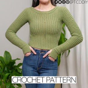 Puede incluir: Un suéter de crochet verde con mangas largas y cuello redondo. El suéter es corto y tiene una textura acanalada. La modelo lleva el suéter con jeans azules.