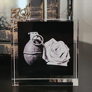 May include: A black and white image of a hand grenade and a rose is encased in a clear, rectangular block. The grenade is on the left, and the rose is on the right. The background is black.