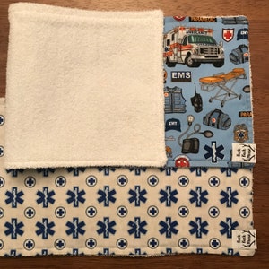 K&ouml;nnte beinhalten: Ein Set aus zwei blauen und wei&szlig;en Stoffquadraten mit einer wei&szlig;en Frottee-R&uuml;ckseite. Ein Quadrat hat einen blauen Hintergrund mit einem sich wiederholenden Muster aus medizinischen Symbolen, einem Krankenwagen und einer Sanit&auml;terweste. Das andere Quadrat hat einen wei&szlig;en Hintergrund mit einem sich wiederholenden Muster aus blauen medizinischen Symbolen. Beide Quadrate haben ein kleines blaues und wei&szlig;es Etikett mit dem Text "Buck Moose".