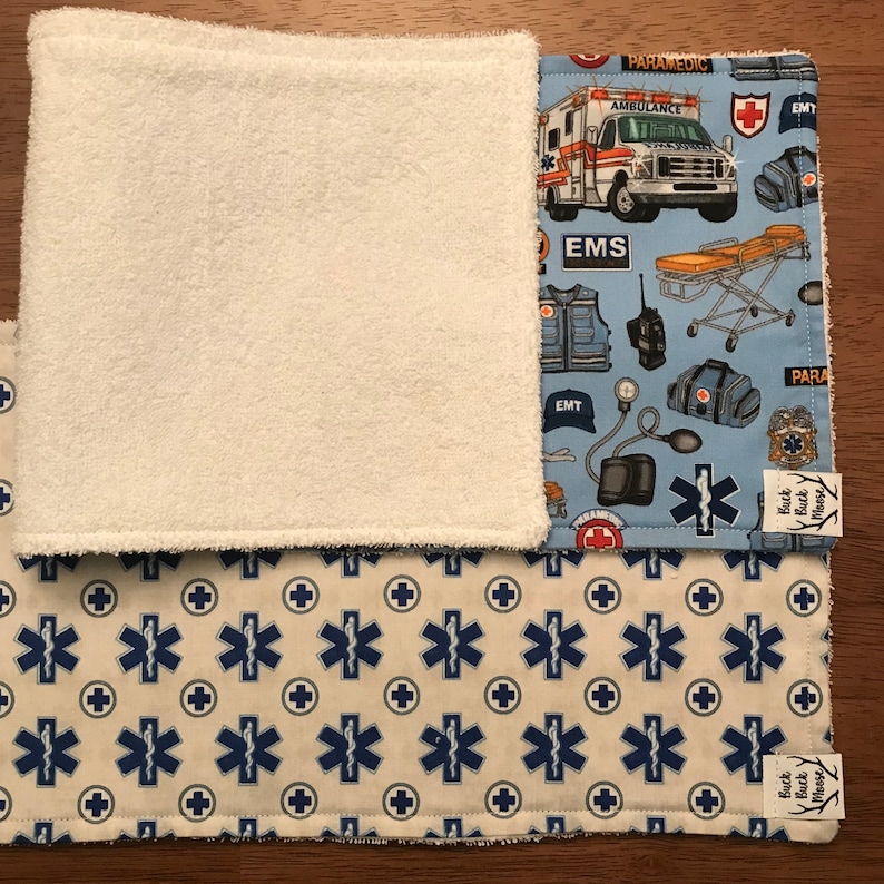 K&ouml;nnte beinhalten: Ein Set aus zwei blauen und wei&szlig;en Stoffquadraten mit einer wei&szlig;en Frottee-R&uuml;ckseite. Ein Quadrat hat einen blauen Hintergrund mit einem sich wiederholenden Muster aus medizinischen Symbolen, einem Krankenwagen und einer Sanit&auml;terweste. Das andere Quadrat hat einen wei&szlig;en Hintergrund mit einem sich wiederholenden Muster aus blauen medizinischen Symbolen. Beide Quadrate haben ein kleines blaues und wei&szlig;es Etikett mit dem Text "Buck Moose".