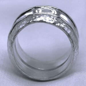 Op de afbeelding: Een zilveren ring met een gehamerde textuur op de buitenste band en een gladde, gepolijste binnenband. De ring heeft een breed bandontwerp en een helder, reflecterend oppervlak. De ring is een sieraad.