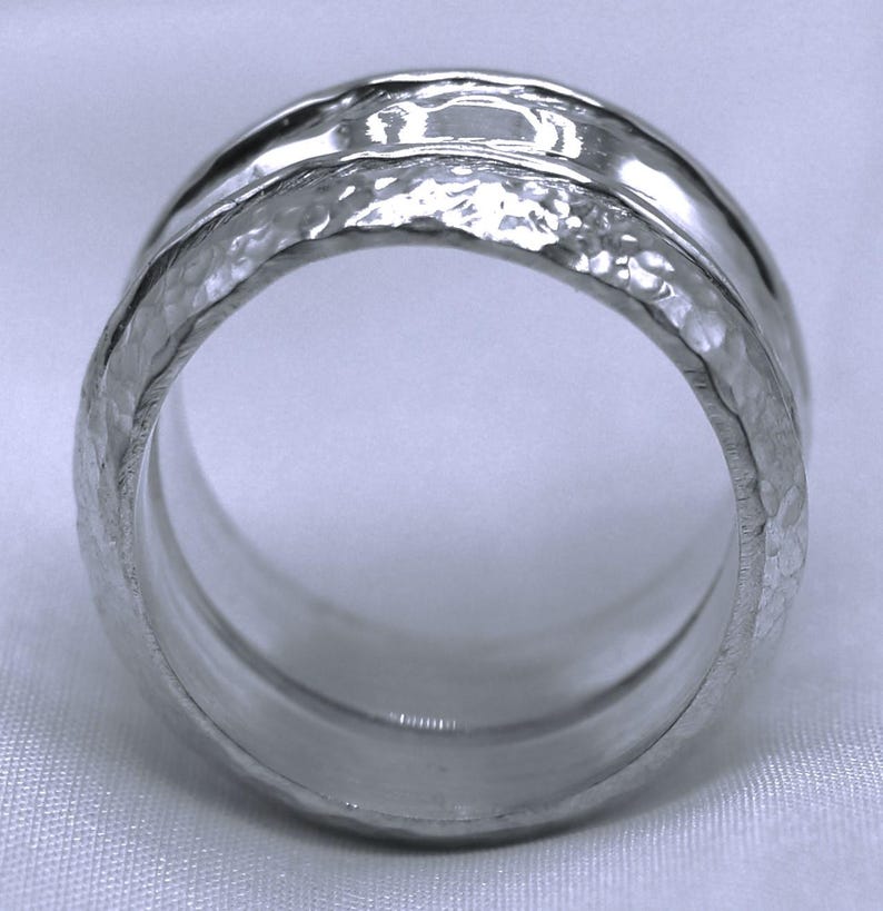 Op de afbeelding: Een zilveren ring met een gehamerde textuur op de buitenste band en een gladde, gepolijste binnenband. De ring heeft een breed bandontwerp en een helder, reflecterend oppervlak. De ring is een sieraad.