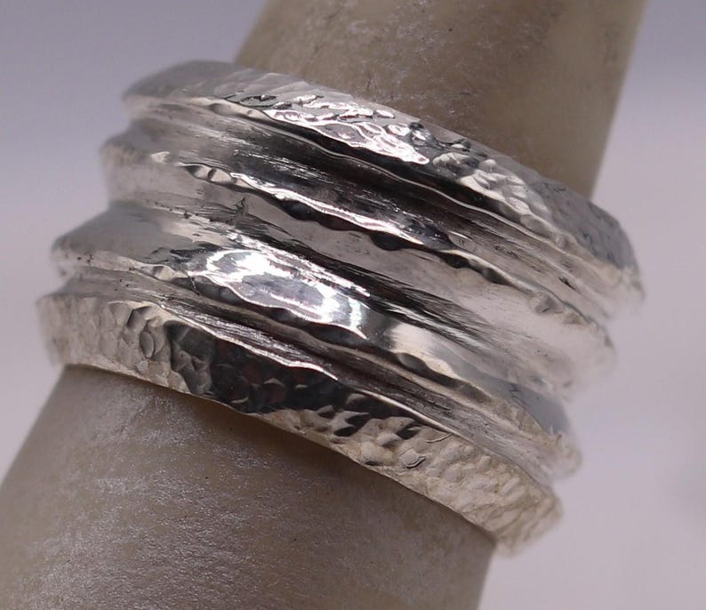 Op de afbeelding: Een zilveren ring met een getextureerde, gehamerde afwerking. De ring heeft een gestapeld ontwerp met meerdere banden, waardoor een gelaagd effect ontstaat. Het metaal reflecteert licht en toont de ingewikkelde details en het vakmanschap van de ring.