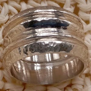Op de afbeelding: Een zilveren ring met een getextureerde, gehamerde afwerking. De ring heeft een brede band met meerdere ribbels en een gepolijst interieur. De ring is geplaatst tegen een cr&egrave;mekleurige, getextureerde achtergrond.