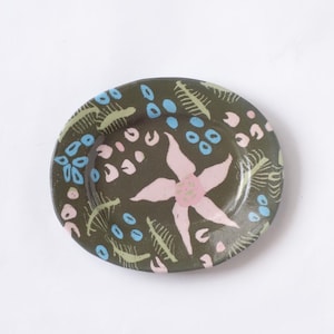 May include: A small, round ceramic plate with a dark olive green background. The plate is decorated with a large pink flower in the center, surrounded by blue and pink floral and leaf patterns. The plate is a decorative item.