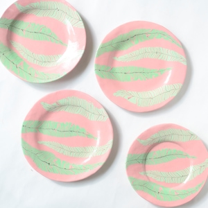 May include: Four pink ceramic plates with a green leaf pattern. The plates are round and have a glossy finish. The leaf design is repeated across the plates, creating a cohesive set. The plates are arranged on a white surface.