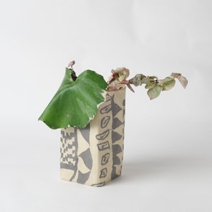 May include: A rectangular vase with a beige background and gray geometric patterns. A large green leaf and a branch with small pink and green leaves are placed inside the vase. The vase is on a white background.