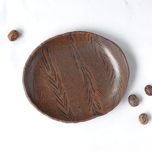 May include: A handmade, brown ceramic plate with a unique, textured surface and a dark, linear pattern. The plate has an organic, irregular shape. Several small, round, brown seeds are scattered around the plate on a white background.