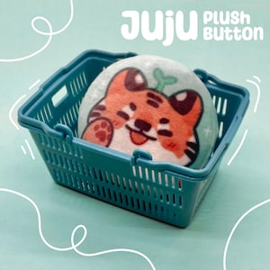 Pode incluir: Um cesto de compras de plástico turquesa contém um botão de pelúcia com o rosto de um tigre de desenho animado. O botão é azul claro com um desenho de tigre laranja e castanho. O texto "JUJU PLUSH BUTTON" é visível.