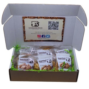Peut inclure: Une boîte en carton marron avec un couvercle blanc est ouverte, révélant une boîte-cadeau remplie de petits sachets de pralines aux noix de pécan. Les sachets sont étiquetés "Sugarland Pecan Orchard" et présentent un logo d'écureuil. La boîte est remplie de papier déchiqueté vert.