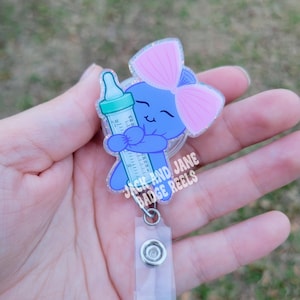 May include: A blue badge reel featuring a cartoon bear holding a baby bottle. The bear has a pink bow and is holding a green and clear bottle. The badge reel has the text "JACK AND JANE BADGE REELS".