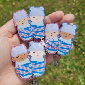 May include: A collection of baby-themed badge reels. Each reel features a sleeping baby design wrapped in a blue and pink striped blanket. The babies wear matching hats or bows. The edges of the badges are outlined with glitter.