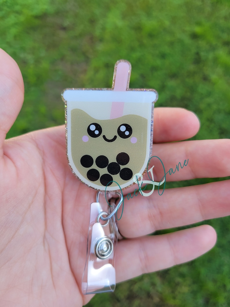 May include: A gold glitter bubble tea charm with a pink straw and black boba pearls. The charm has a cute cartoon face with big eyes and a smile. It is attached to a clear plastic retractable badge holder.