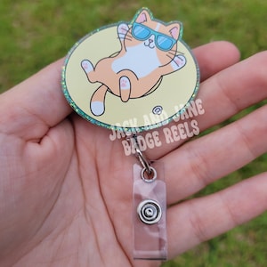 May include: A badge reel featuring a cartoon cat wearing sunglasses, relaxing on a yellow circle with a glittery border. The reel has a clear plastic retractable cord and a metal clip. Text on the badge reel reads "JACK AND JANE BADGE REELS".