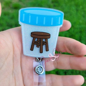 May include: A white glitter badge reel with a blue lid and a brown stool inside. The reel has a clear plastic strap and a silver swivel clip.