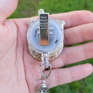 May include: Gold glitter retractable badge holder with a clear plastic case and a silver clip. The holder is attached to a clear plastic strap with a silver swivel clip.