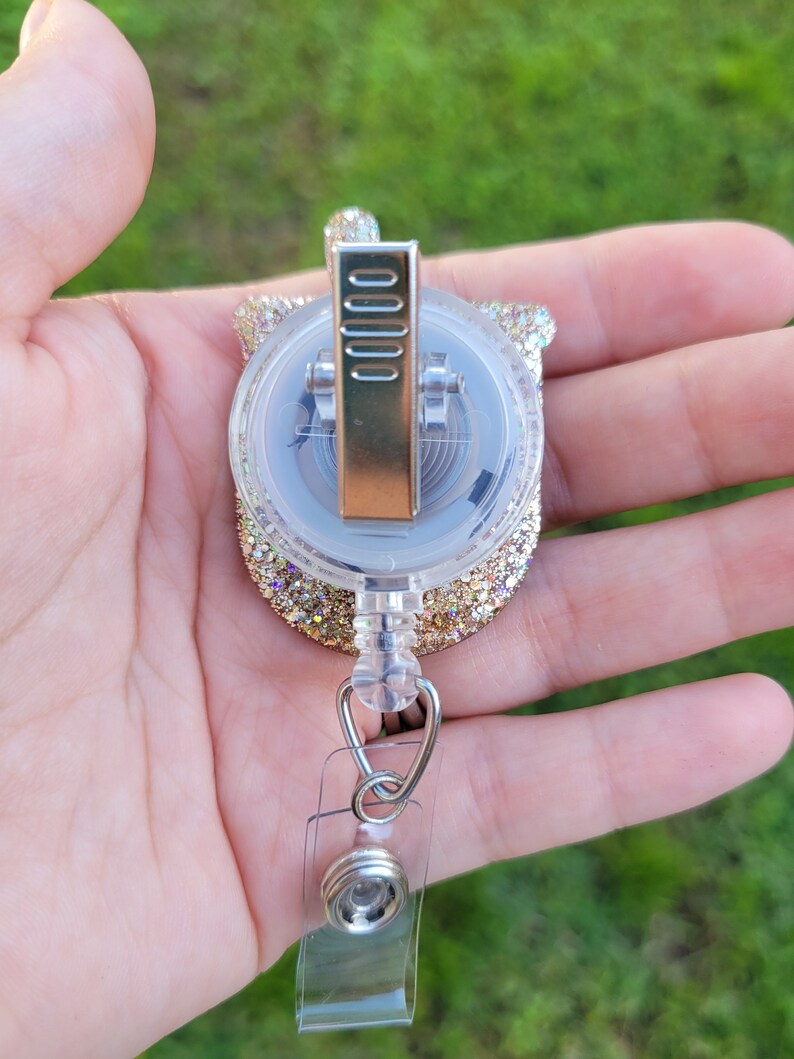 May include: Gold glitter retractable badge holder with a clear plastic case and a silver clip. The holder is attached to a clear plastic strap with a silver swivel clip.