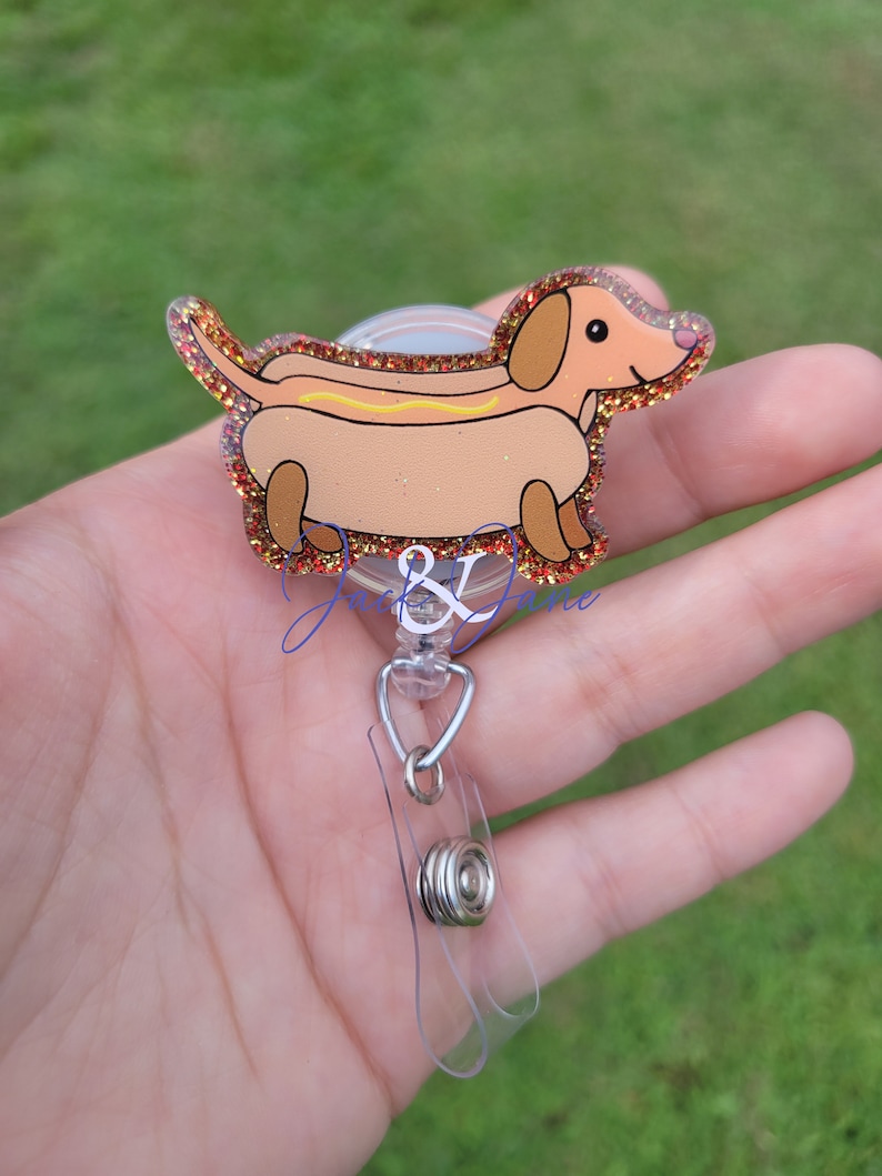 May include: A glitter-covered dachshund shaped badge reel with a hot dog design. The dachshund is brown with a yellow hot dog and mustard. The reel is clear plastic with a silver retractable cord.
