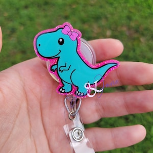 May include: A turquoise glitter dinosaur with a pink bow on its head attached to a clear retractable badge holder.