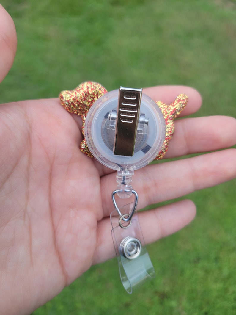 May include: A gold and red glitter dog charm attached to a clear retractable badge holder with a silver clip.