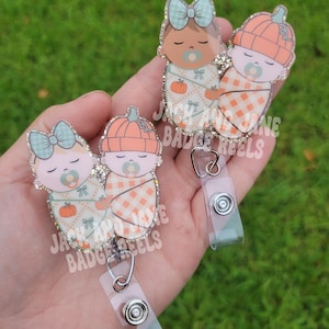 May include: Two glittery acrylic badge reels featuring two swaddled babies, one with a light blue bow and pumpkin-patterned blanket, the other with an orange pumpkin hat and plaid blanket. Both have pacifiers. Text reads JACK AND JANE BADGE REELS.