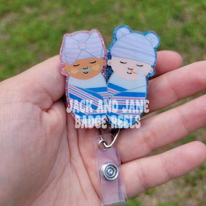 May include: A badge reel featuring two cartoon babies wrapped in blue and pink striped blankets. The babies wear blue and pink hats and have pacifiers. The text "JACK AND JANE BADGE REELS" is printed on the badge reel.