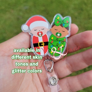 May include: A badge reel featuring two baby designs, one dressed as Santa Claus and the other wrapped in Christmas lights. The text "available in different skin tones and glitter colors" is included. The badge reel has a clear plastic strap and a silver metal clip.