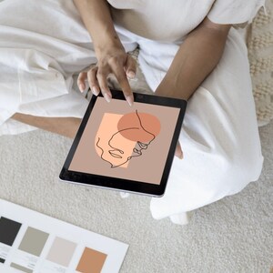 May include: A person is holding a tablet displaying a digital illustration of a face with a peach and brown color scheme. The tablet is on a white surface.